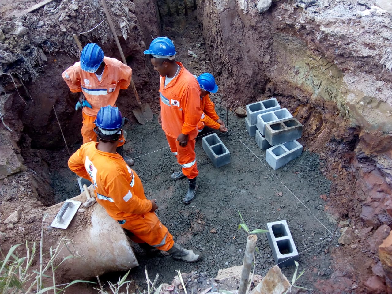 Quatro trabalhadores com uniforme laranja estão dentro de um buraco que é um pouco mais fundo que seus corpos. Um está sentado no tubo de cimento, onde será montada uma galeria. No fundo, massa de concreto. ao lado, blocos de cimento, para fazer a parede.